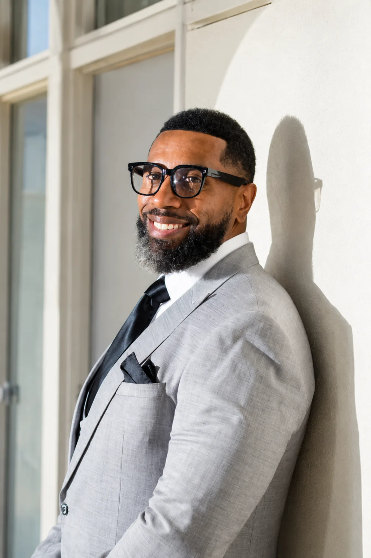 Cory J. Chavis, a sitting principal and school-culture speaker, smiling in a light gray suit, black tie, and glasses, standing in front of a sunlit wall